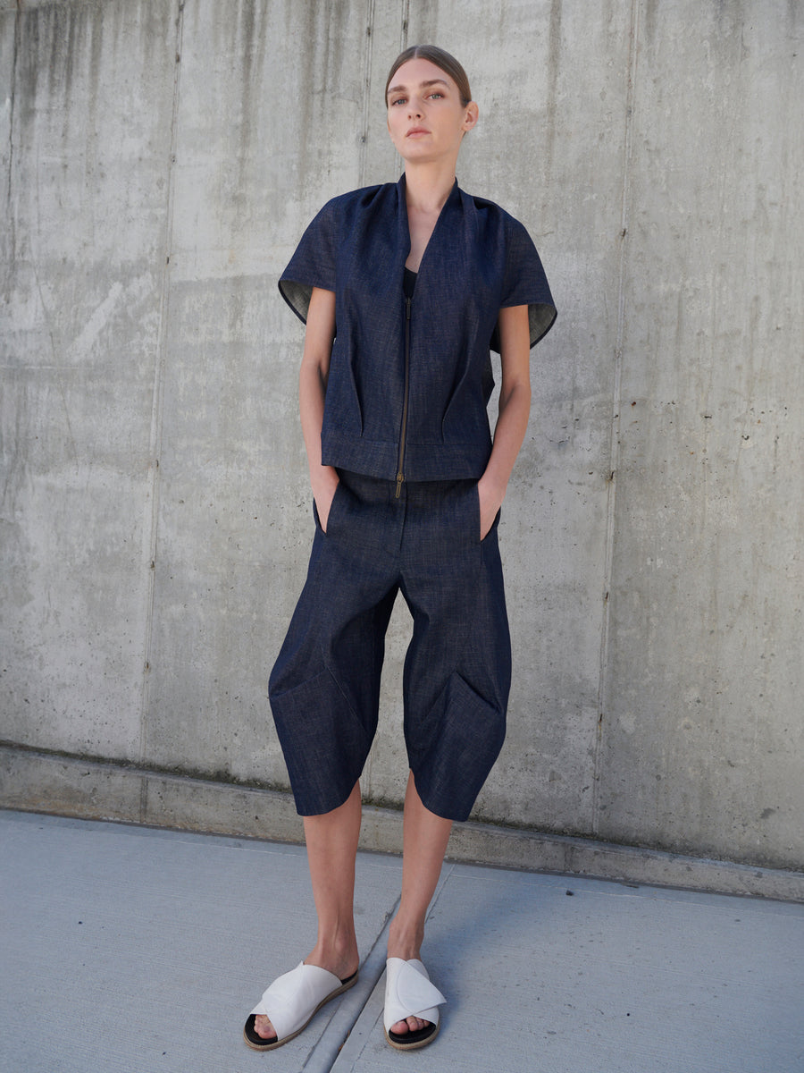 A person stands against a concrete wall wearing a modern outfit by Zero + Maria Cornejo, featuring Beetle Shorts crafted from GOTS-certified cotton with loose short sleeves and wide cropped pants. They have hands in pockets and wear white slide sandals. The background is minimalistic.