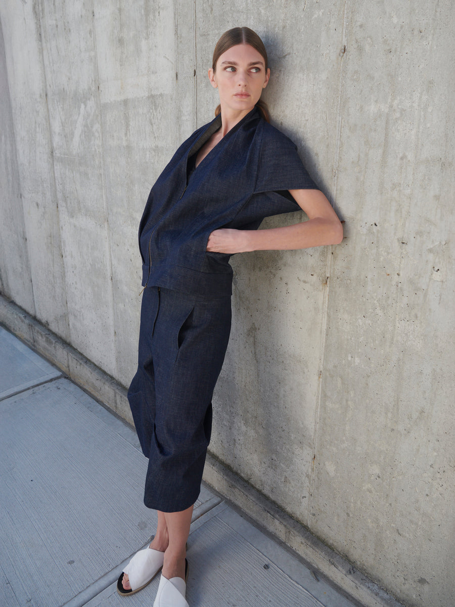 A person stands against a concrete wall wearing a dark blue, oversized jacket with matching pants. Their light hair is pulled back as they look to the side. They are wearing white slip-on shoes and Zero + Maria Cornejo's Beetle Shorts made from organic denim.