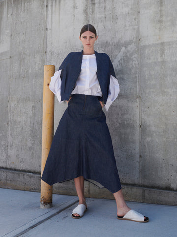 A person stands against a concrete wall wearing a sustainable fashion ensemble featuring the Aissa Joi Skirt by Zero + Maria Cornejo with an asymmetrical hem, paired with a structured jacket and white blouse. They complete the look with white slide sandals. A yellow pole is nearby.