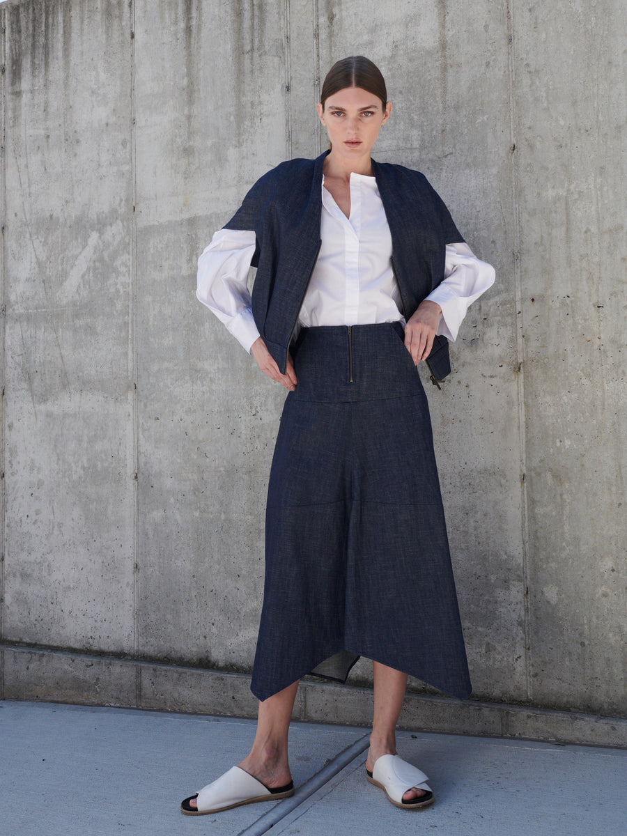 A person confidently poses against a concrete wall in a dark blue organic denim outfit, paired with a white shirt and white slide sandals. Sporting short hair and hands on hips, they wear the Zero + Maria Cornejo Aissa Joi Skirt with an asymmetrical hem, embodying sustainable fashion.