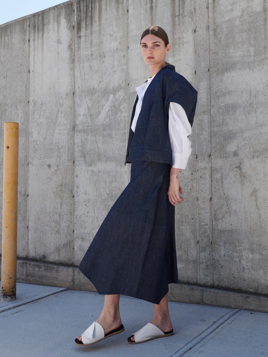 A person with pulled-back hair stands against a concrete wall wearing a dark blue sleeveless vest, the Aissa Joi Skirt by Zero + Maria Cornejo over a white shirt. They pair this look with white sandals and gaze at the camera, embodying sustainable fashion with style.