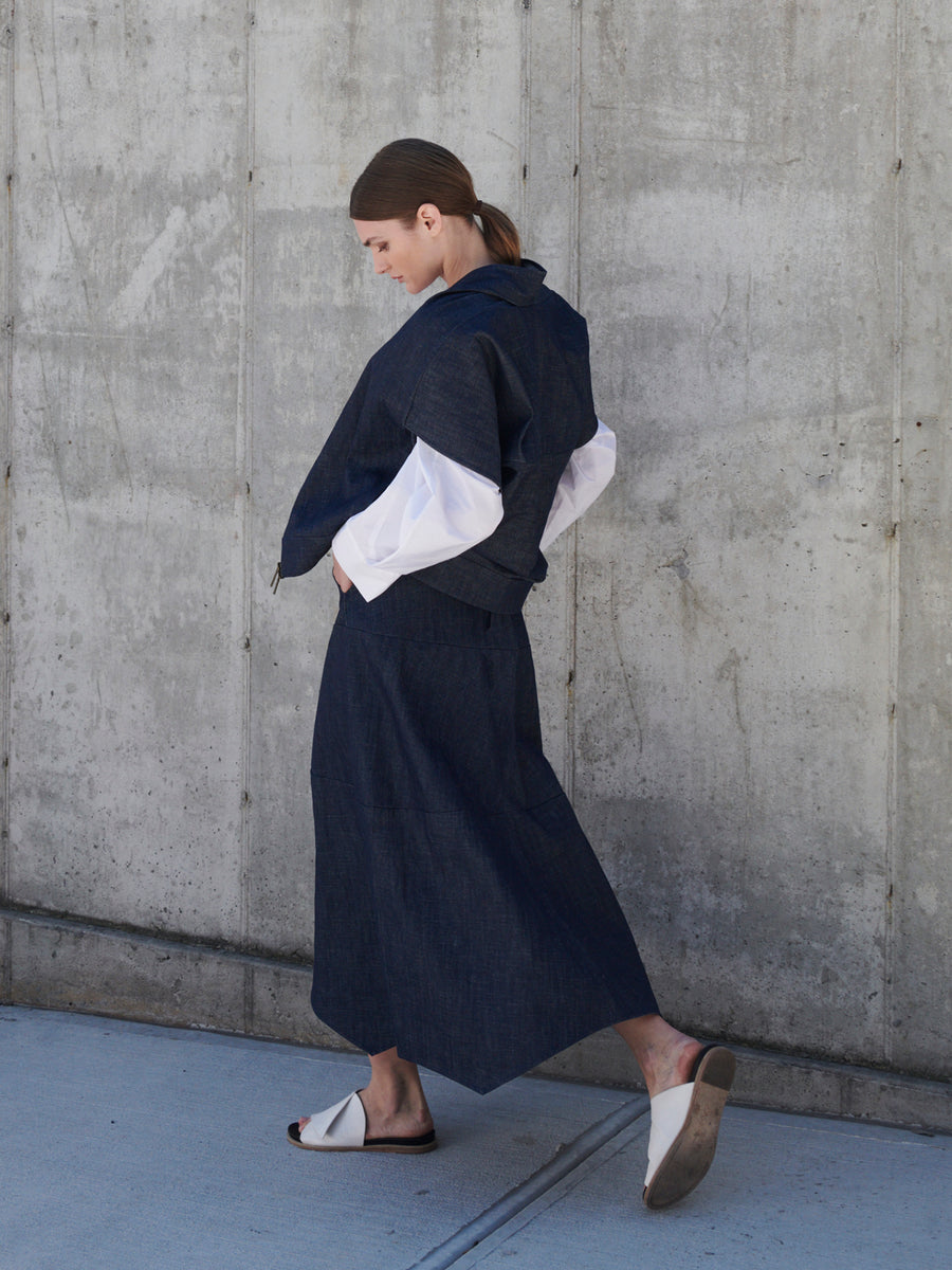 A person with brown hair tied back is wearing a dark blue jacket and white shirt with wide sleeves. Their Aissa Joi Skirt, by Zero + Maria Cornejo, is long and made of organic denim. They walk against a concrete wall, looking down, in white sandals, embodying sustainable fashion.