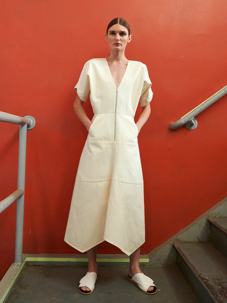 A person in the Aissa Joi Dress by Zero + Maria Cornejo, a GOTS-certified off-white V-neck dress with an asymmetrical hem, stands by the red wall and metal railings on the staircase. With matching slip-on sandals and hands in pockets, they effortlessly blend style with sustainability.