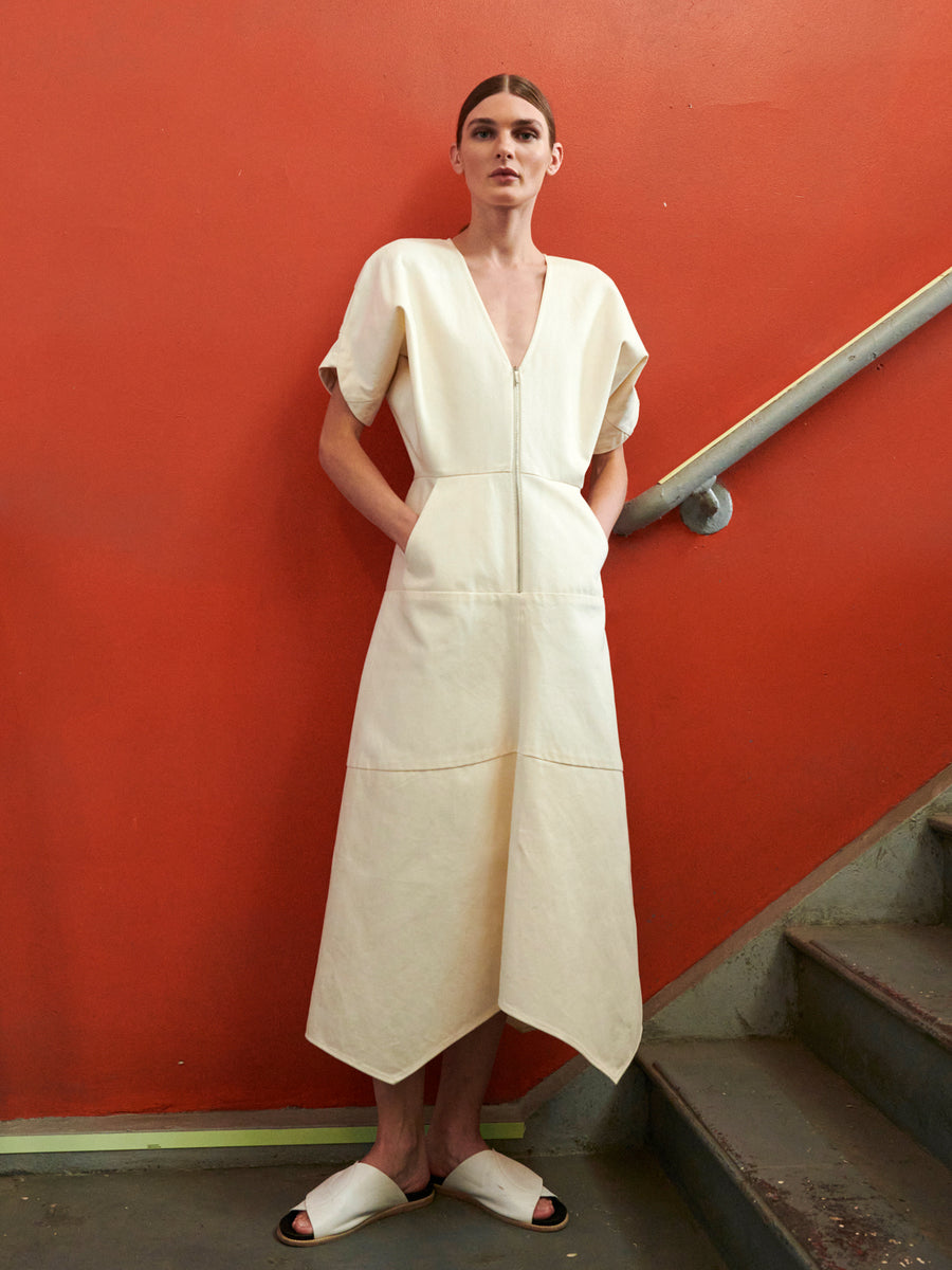 On a staircase against a vibrant orange wall, a person with dark hair in open-toed sandals wears the Aissa Joi Dress by Zero + Maria Cornejo. This cream-colored dress, made of GOTS-certified cotton, features a deep V-neck, wide short sleeves, and front pockets.