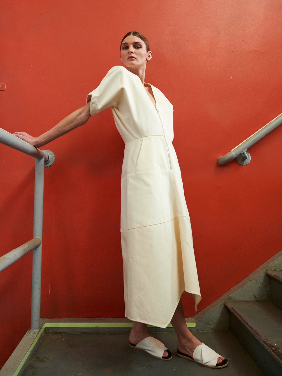 A person in a cream Aissa Joi Dress by Zero + Maria Cornejo leans against a staircase railing before an orange wall, wearing white sandals. With short hair, they look upward with confidence.