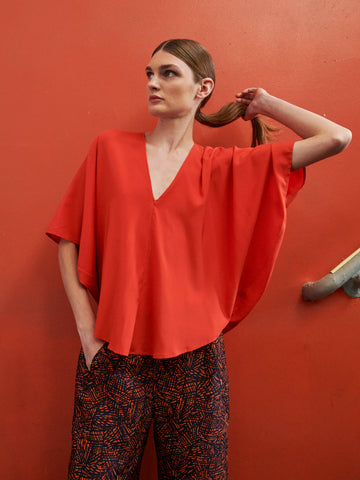 A person poses against an orange wall, wearing Zero + Maria Cornejo's Circle Top and patterned pants. The silk fabric of the top shimmers as they hold their ponytail with one hand while placing the other in their pocket, gazing slightly upward.