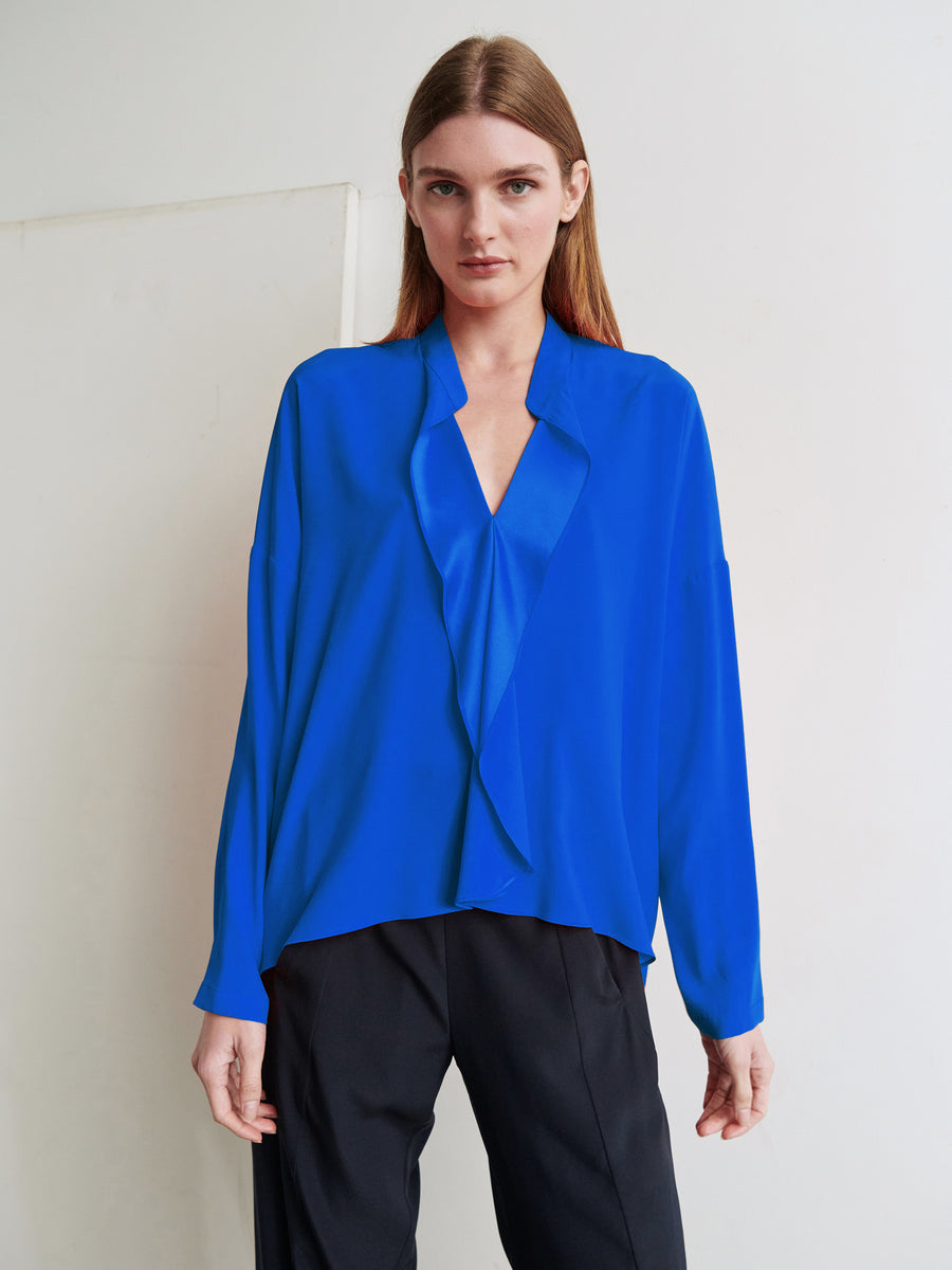 A person with long brown hair stands against a white wall wearing a vibrant blue Long-Sleeved Fin Gaban Shirt from Zero + Maria Cornejo, paired with loose black pants. They look directly at the camera with a neutral expression.