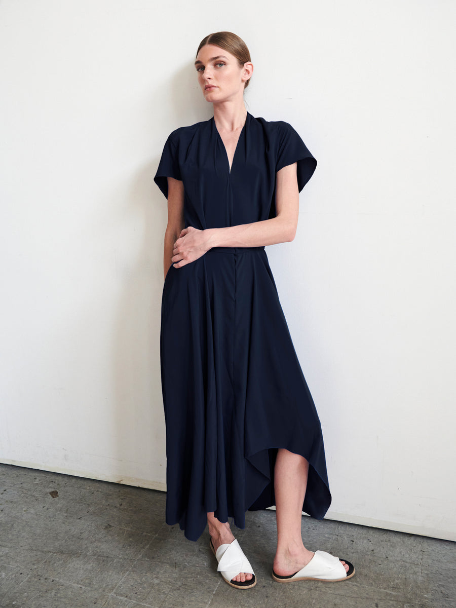 A person stands against a white wall, deep in thought, wearing the navy blue Maki Wave Dress by Zero + Maria Cornejo. The flowy charmeuse design with an asymmetrical hemline is paired with white slide sandals. Beneath them lies a gray concrete floor, adding an urban touch to the serene scene.