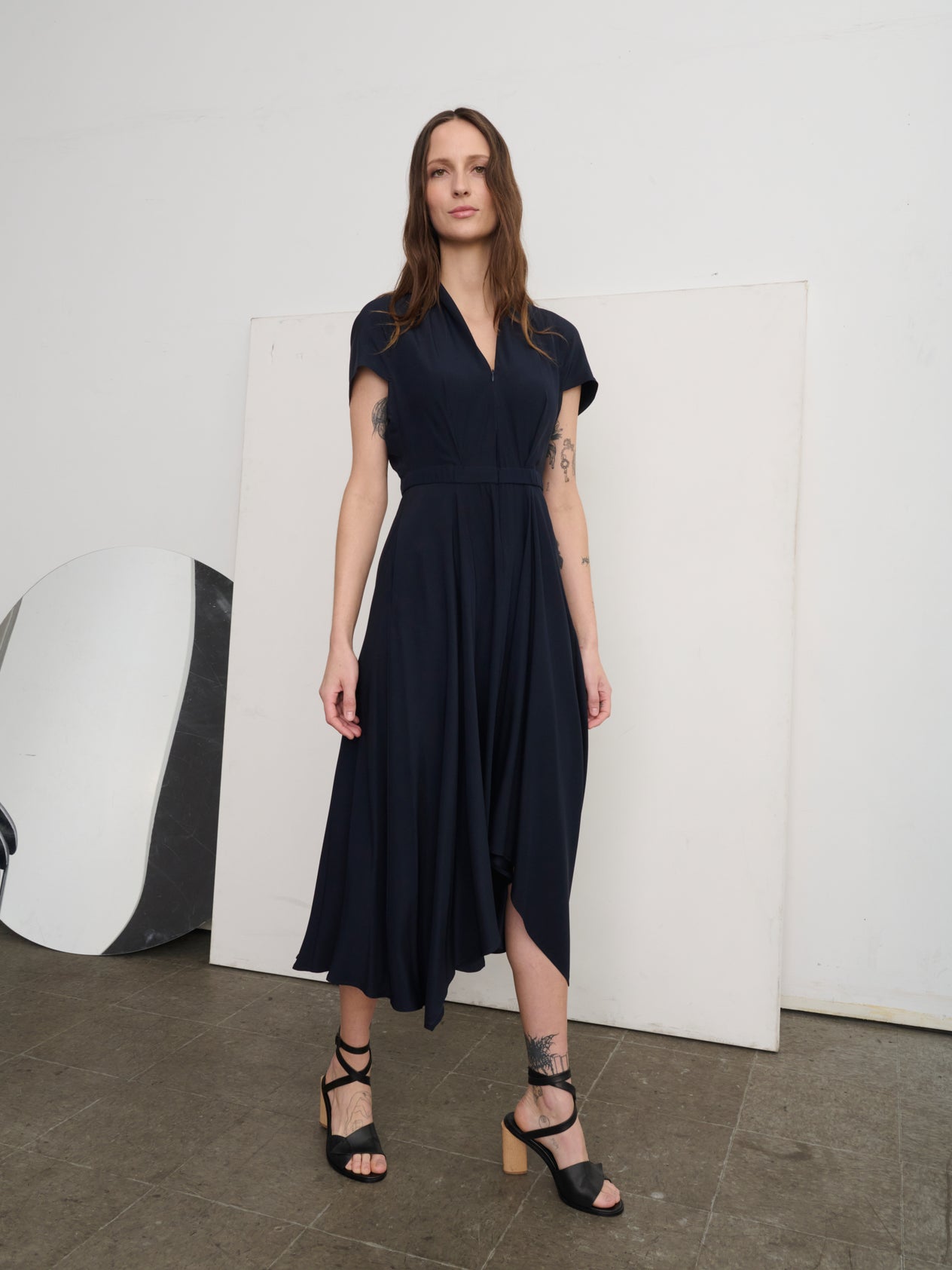 A woman stands against a white wall wearing the vibrant blue Maki Wave Dress by Zero + Maria Cornejo, made of stretch silk charmeuse, with a short sleeve and asymmetrical hemline. She completes her look with white slide sandals and her hair pulled back, gently resting one hand near her neck.