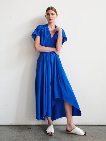 A woman stands against a white wall wearing the vibrant blue Maki Wave Dress by Zero + Maria Cornejo, made of stretch silk charmeuse, with a short sleeve and asymmetrical hemline. She completes her look with white slide sandals and her hair pulled back, gently resting one hand near her neck.