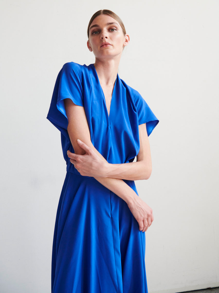 A person poses against a plain background, wearing the Maki Wave Dress by Zero + Maria Cornejo. The blue silk charmeuse dress features short sleeves and a V-neck. They have their hair pulled back and pose with one arm across the body, holding the opposite elbow.