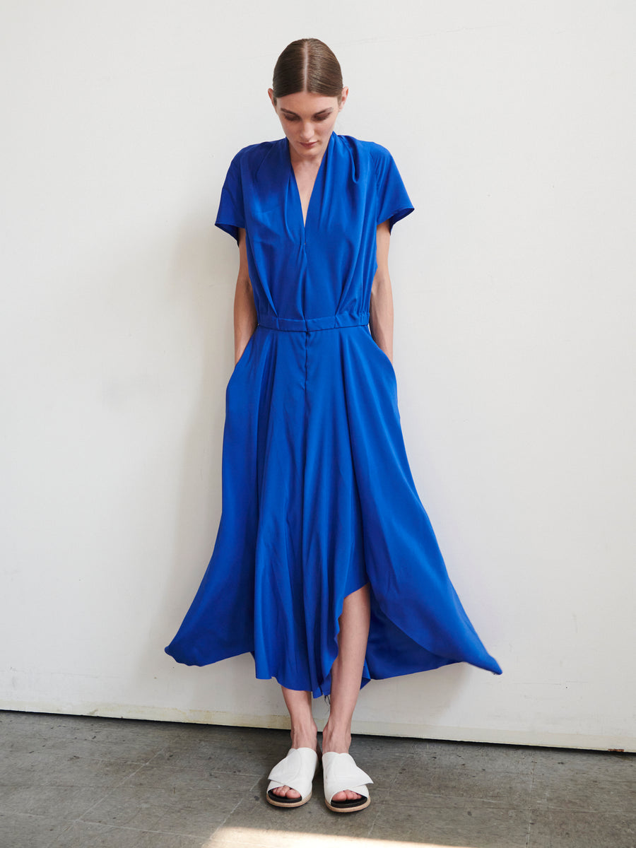 A person with slicked-back hair stands against a plain white wall, wearing the Maki Wave Dress by Zero + Maria Cornejo—a flowing blue V-neck dress with an asymmetrical hemline and short sleeves—paired with white sandals. They have their hands in the pockets, looking downwards.