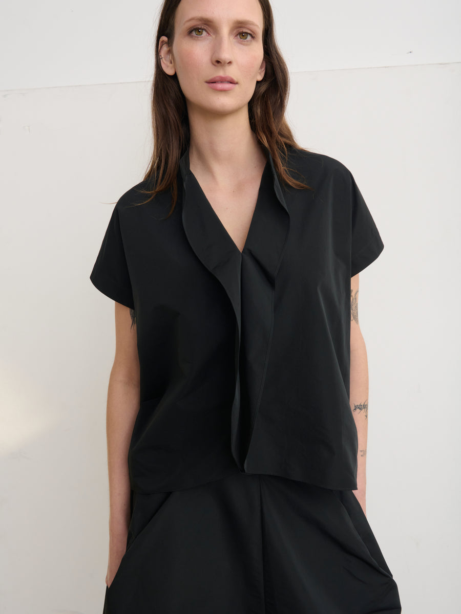 A woman with long brown hair wears the loose-fitting black Fin Gaban Shirt by Zero + Maria Cornejo. She stands against a light background, looking directly at the camera with a neutral expression, hands in pockets.