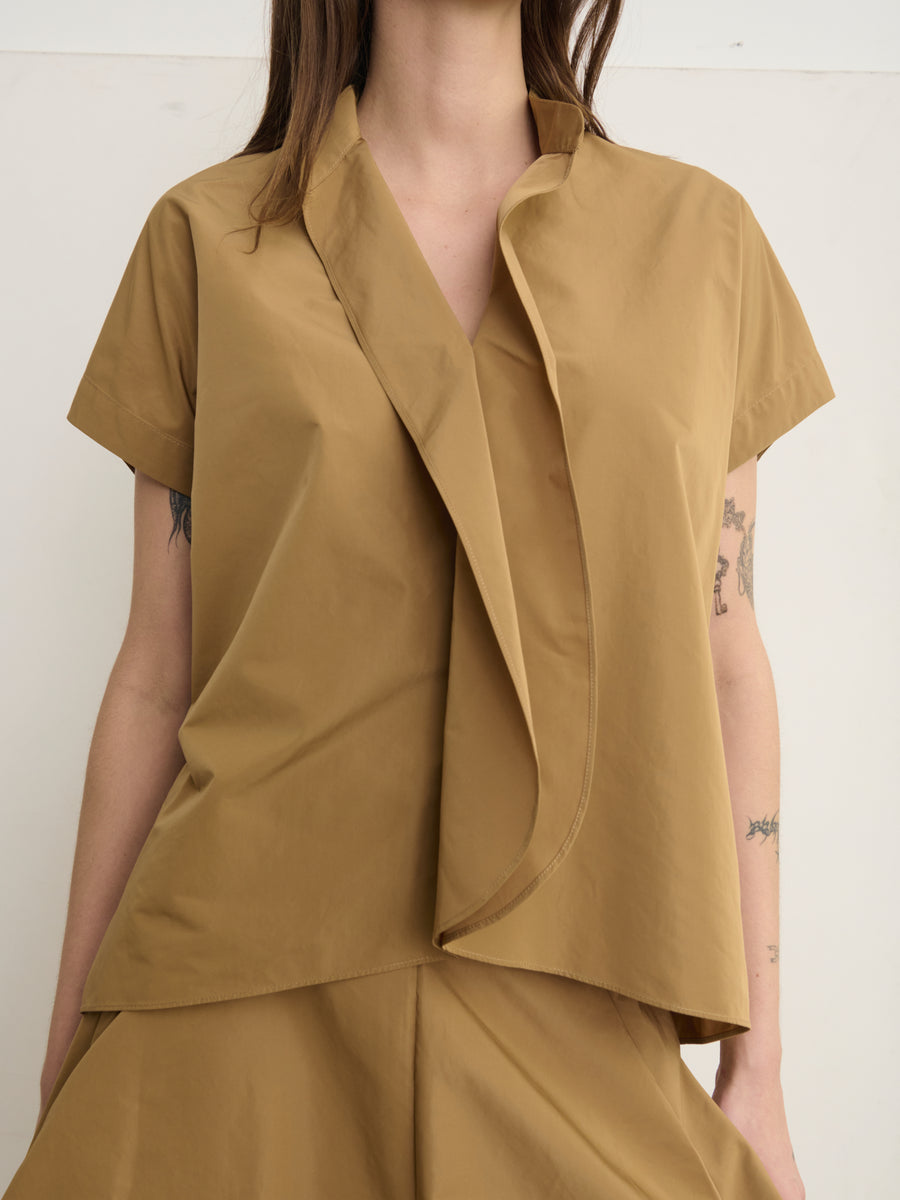 A person wears the Fin Gaban Shirt by Zero + Maria Cornejo, a stylish, light brown, short-sleeved blouse with an asymmetrical draped collar. Crafted from cotton shirting and paired with matching pants, it highlights modern minimalism. Tattoos are visible on the arms, while only the upper body is shown.