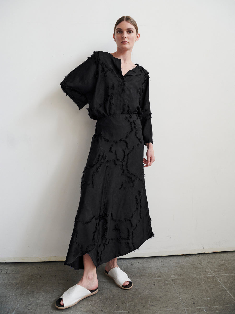 A person with hair tied back wears a textured black long-sleeve top and Circle Skirt by Zero + Maria Cornejo made from FSC viscose. They pose against a plain white wall in white open-toe sandals, one hand in their pocket.