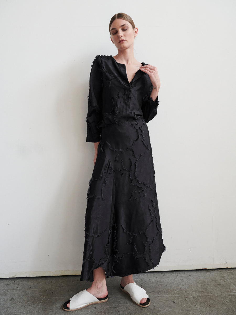 A woman stands barefoot against a plain white wall, wearing the Zero + Maria Cornejo Circle Skirt. Her hair is tied back as she softly gazes downward, showcasing her chic yet sustainable style, with one foot peeking forward in white open-toe slippers.