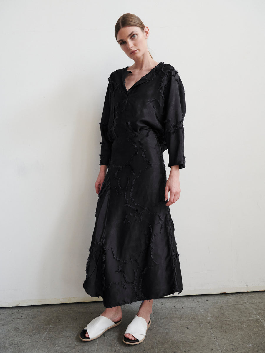 The person wears a long-sleeved black dress with textured jacquard fabric from Zero + Maria Cornejo, standing against a plain wall. They have straight light brown hair and wear white slide sandals. The concrete floor elegantly contrasts with the outfit, highlighting its unique texture.