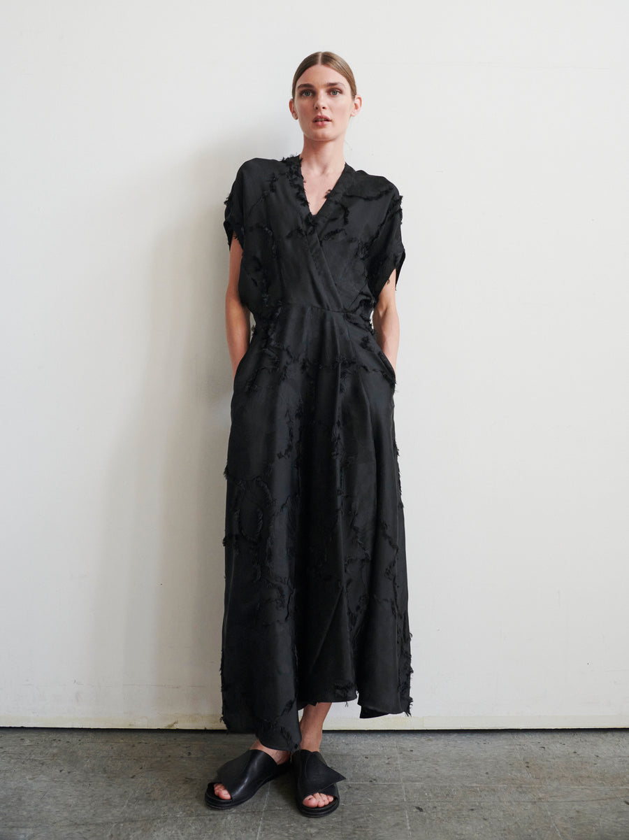 Dressed in Zero + Maria Cornejo's Long Aki Wave Dress with its textured jacquard fabric, asymmetrical skirt, short sleeves, and pockets, a person stands against a plain white wall. Black slip-on sandals complete their look on the simple concrete floor.