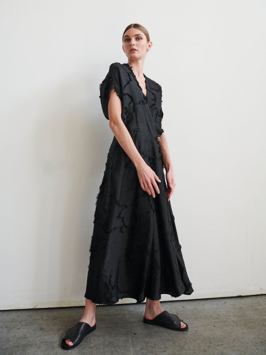 A person stands against a white wall wearing the Long Aki Wave Dress by Zero + Maria Cornejo. It features short sleeves, a V-neckline, jacquard fabric, and textured details. They pair it with black sandals and have short, styled hair.