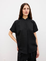 A woman with long brown hair stands against a white background, wearing the Mare Shirt in black by Zero + Maria Cornejo, a short-sleeved, loose-fitting top. She pairs it with black pants and has a neutral expression with one hand on her hip.
