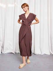 A person stands before white curtains wearing the Zero + Maria Cornejo Mido Dress in Chocolate, a short-sleeved V-neck wrap style made from lightweight woven fabric, paired with beige slide sandals and hands resting on their lower back.