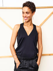 A woman smiles in the Ibit Top by Zero + Maria Cornejo paired with black pants. With short curly hair, dangling earrings, and a bracelet, she poses with her left hand in her pocket against a modern light background.