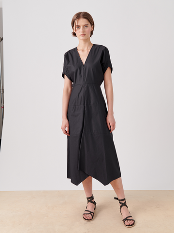 A woman with short brown hair wears a black, short-sleeved, V-neck Aissa Joi Dress from Zero + Maria Cornejo with an asymmetrical hem, standing on a light-colored wooden floor against a white background. She pairs the dress with black strappy sandals and faces the camera with a calm expression.