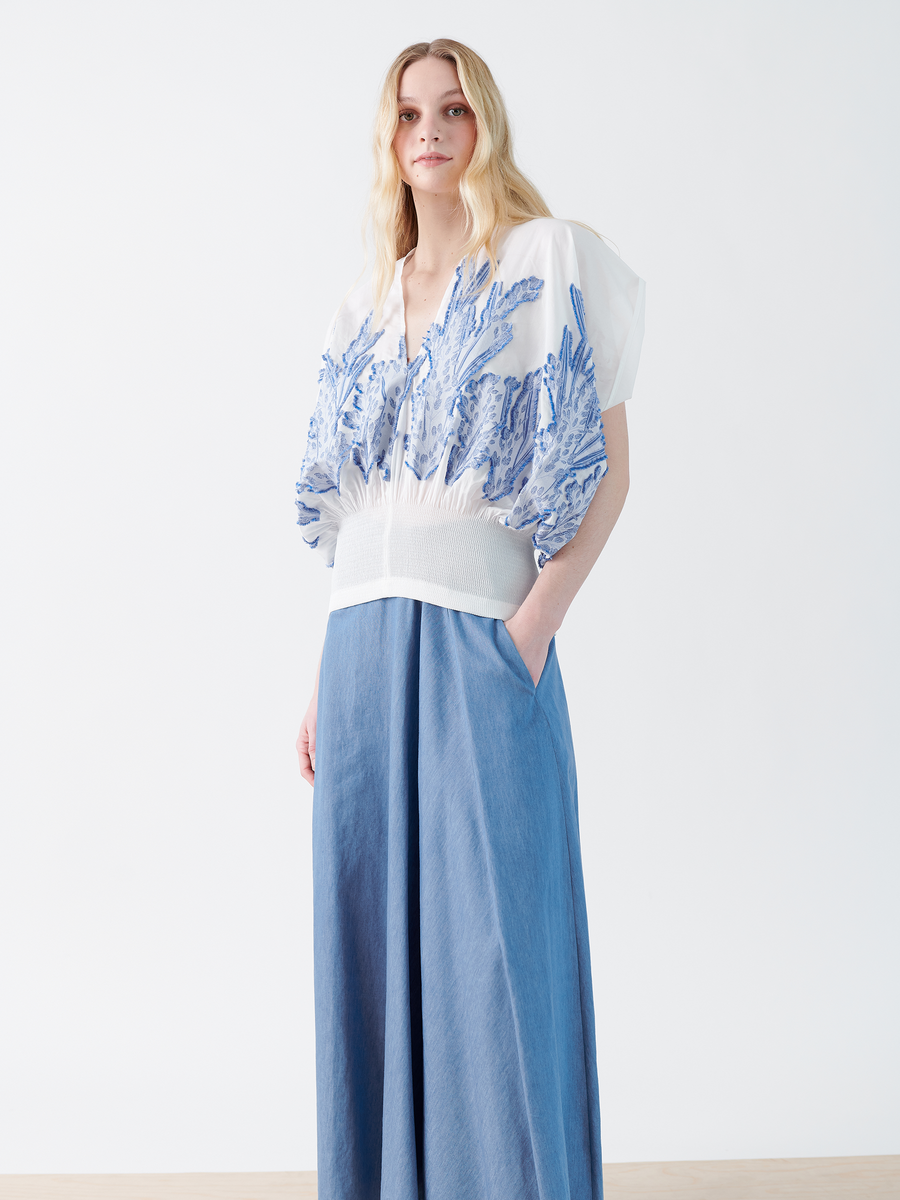 A woman with long blonde hair wears Zero + Maria Cornejo's cotton-blend white blouse, adorned with blue embroidery and a voluminous design, tucked into the Long Wave Skirt - Final Sale. She casually rests her hands in the skirt pockets against a plain white background.