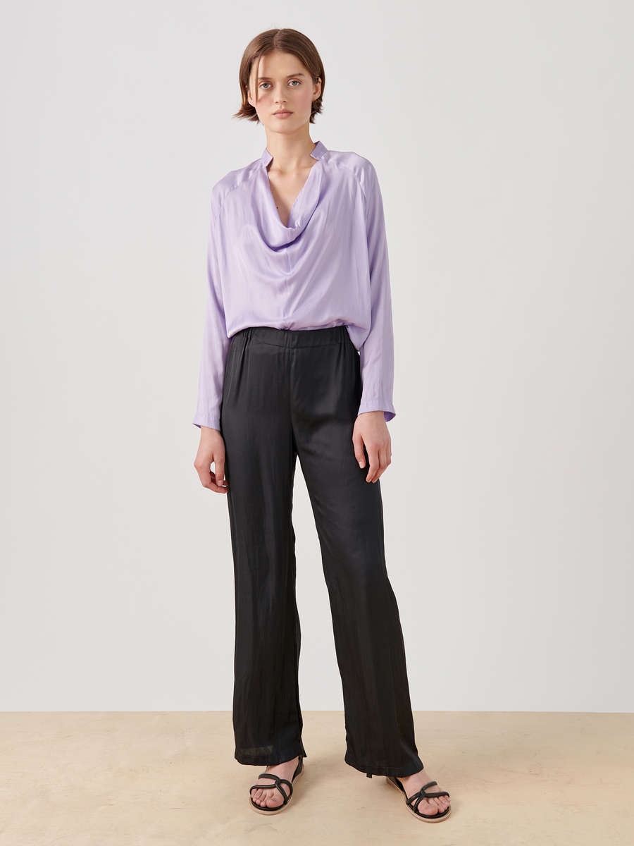 Against a plain background, someone wears the Long-Sleeved Evi Top by Zero + Maria Cornejo, featuring a light purple washed georgette fabric and draped open neckline. They pair it with black wide-leg pants, black sandals, and have simply styled short dark hair.