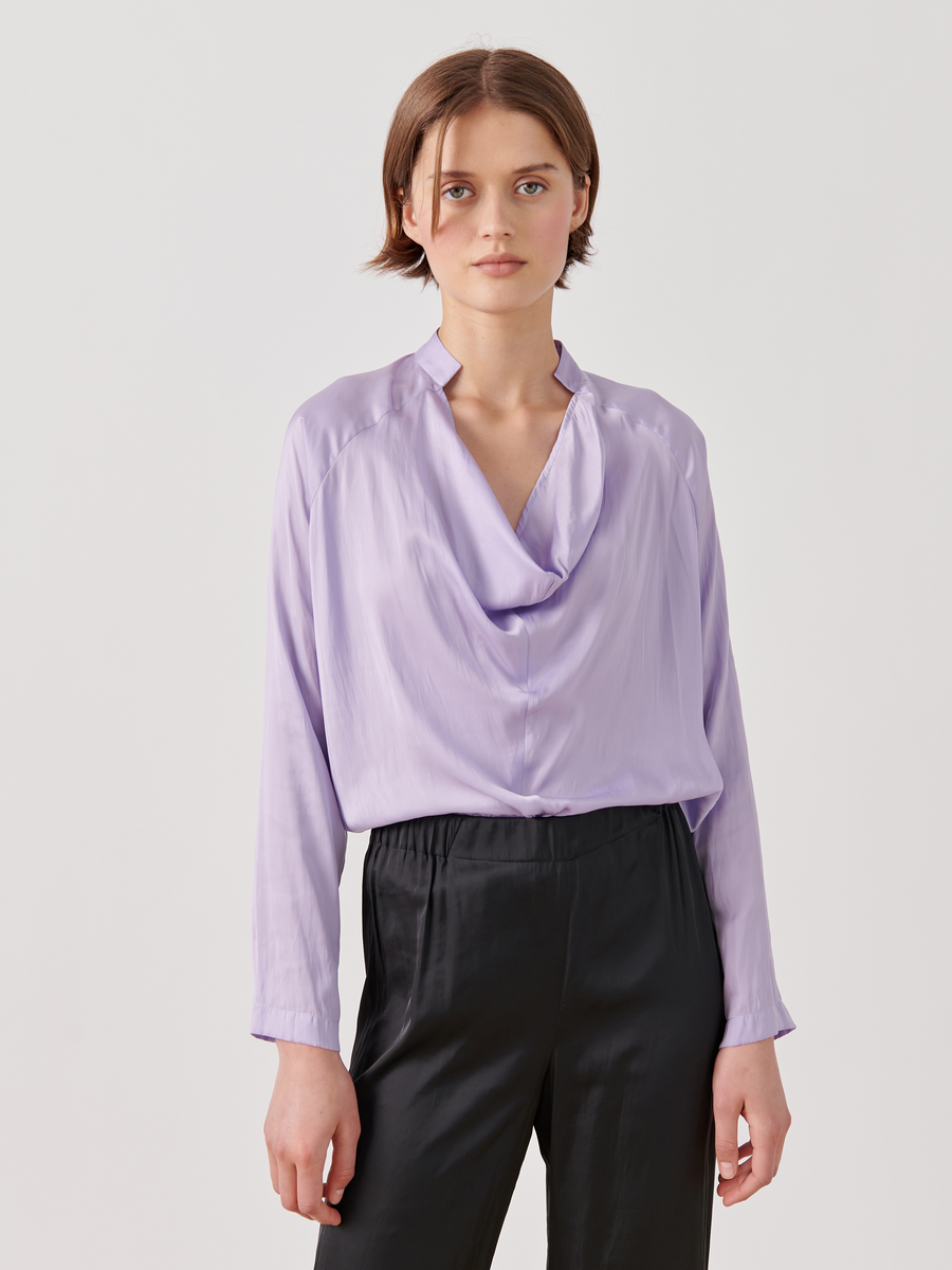 A person is standing against a plain white background wearing a Zero + Maria Cornejo Long-Sleeved Evi Top in lavender washed georgette featuring a draped open neckline, paired with black pants. They have short brown hair and a neutral expression.