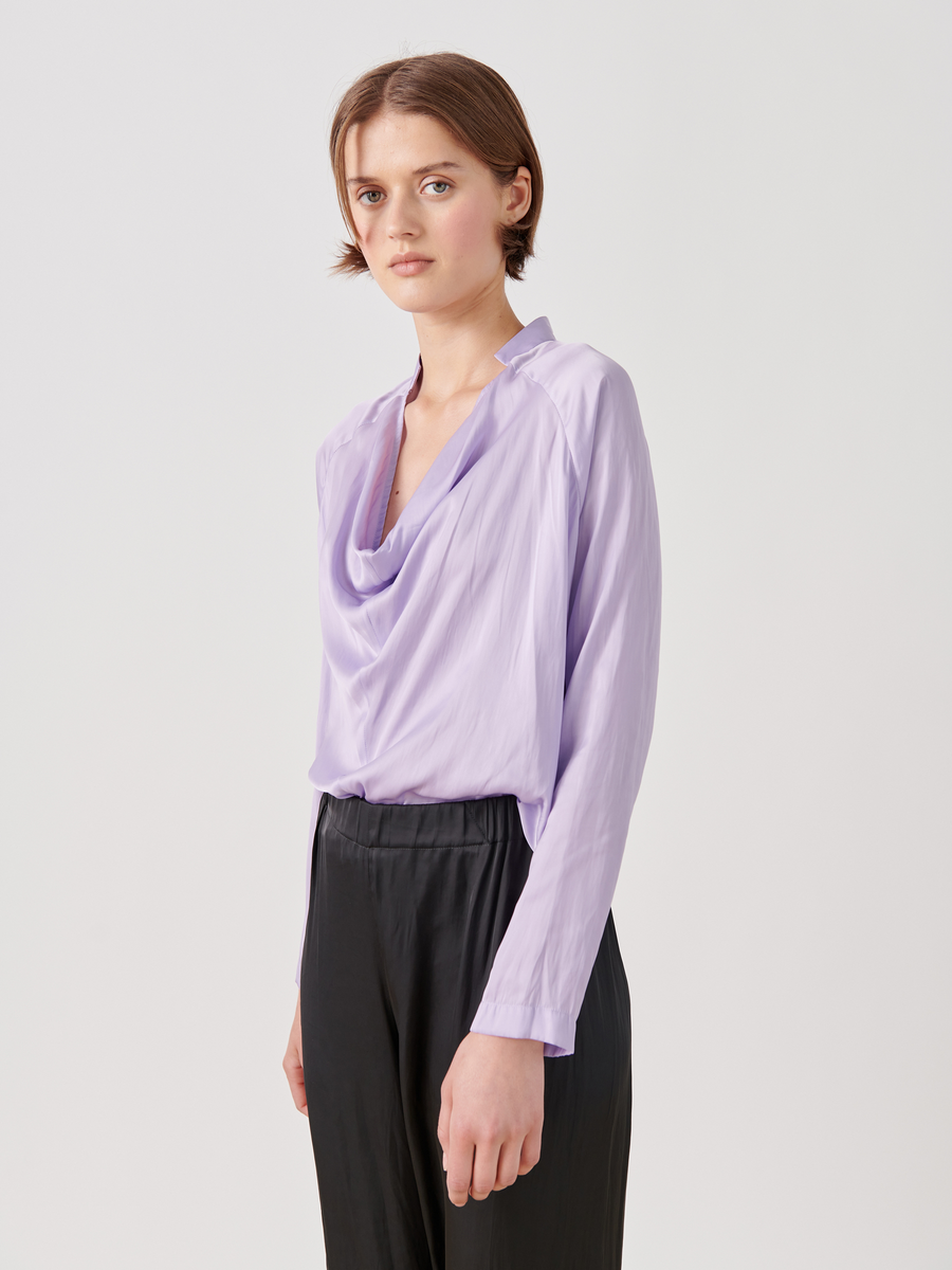 A short brown-haired individual wears the Long-Sleeved Evi Top from Zero + Maria Cornejo, styled with black pants. The light purple washed georgette top features a draped open neckline. They pose against a plain white background, gazing at the camera with a neutral look.