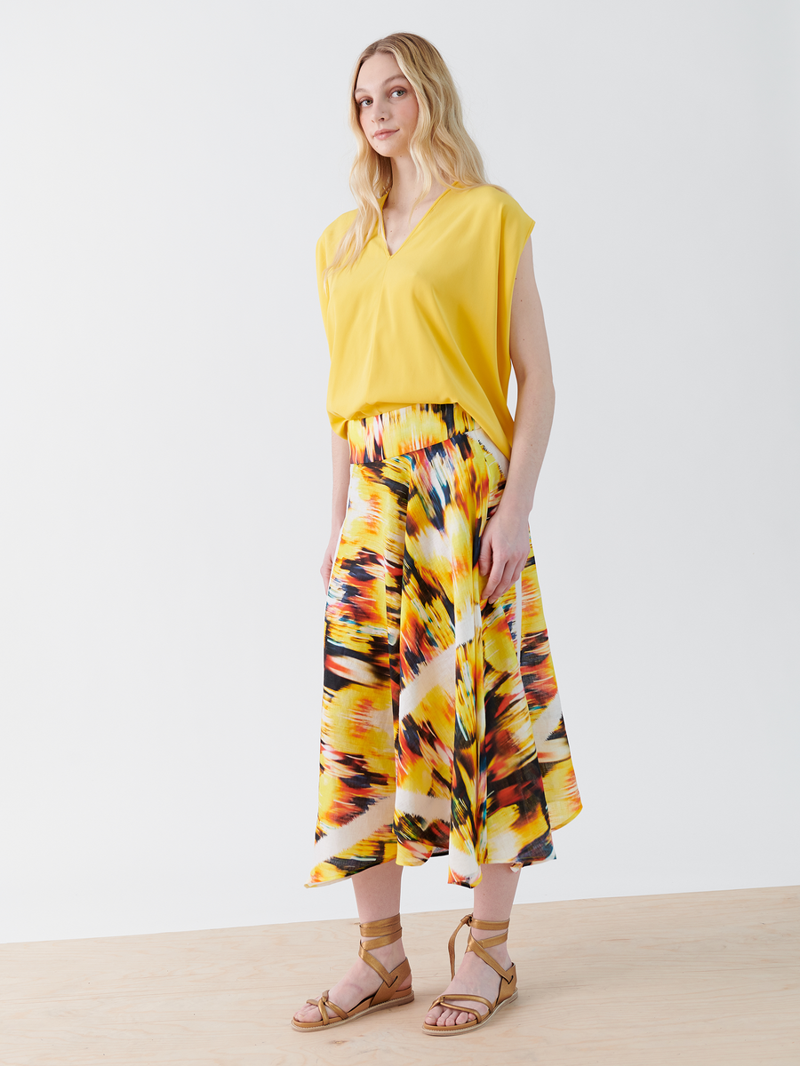 A woman with long blond hair is wearing a sleeveless yellow top and the Aki Wave Skirt by Zero + Maria Cornejo, showcasing shades of yellow, orange, and black. She stands on a light wood floor against a white background, paired with tan sandals.