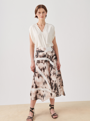 A person with short brown hair stands against a plain background wearing a white V-neck blouse, the Aki Wave Skirt by Zero + Maria Cornejo featuring multicolor abstract designs via digital inkjet printing, and black strappy sandals. They have a neutral expression.