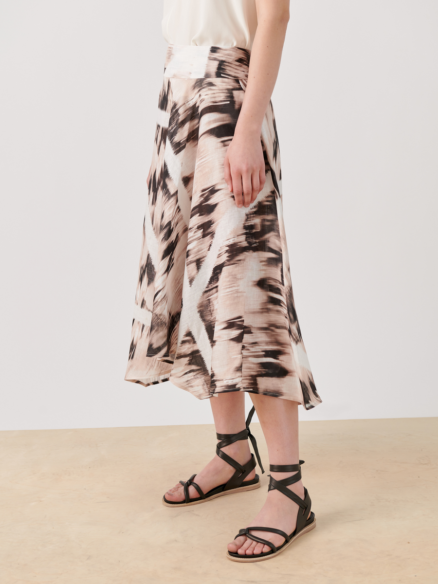 A person is wearing the Aki Wave Skirt by Zero + Maria Cornejo, featuring an asymmetrical, flowy design with abstract brown and white patterns. They pair it with black, strappy sandals against a plain background that highlights the skirt's intricate details.