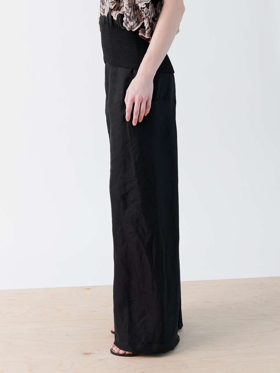 Barefoot on a light wooden floor, someone wears Zero + Maria Cornejo's Wrap Pant, styled with a textured top. The black maxi skirt-inspired look flows gracefully against the plain white background, highlighting comfort and style reminiscent of lightweight linen.