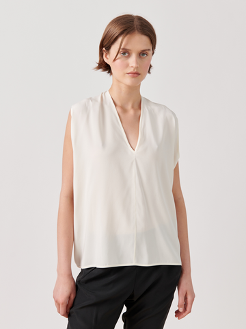 A person with short brown hair, standing against a plain white background, wears a sleeveless V-neck Ina Tank from Zero + Maria Cornejo and black pants. They are looking straight at the camera with a neutral expression.