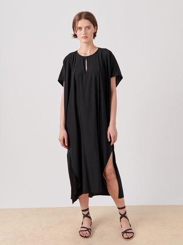 A person wearing a loose-fitting, stretch silk charmeuse Curve Rae Caftan by Zero + Maria Cornejo with short sleeves and a keyhole neckline. The mid-length dress features side slits and falls to mid-calf. Paired with black strappy sandals, the individual stands against a plain, light-colored background.