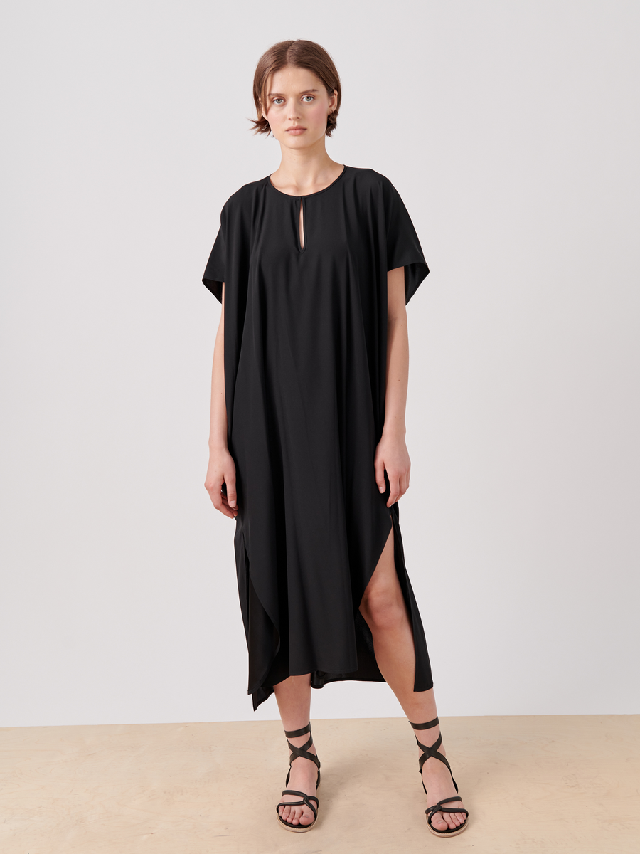 A person wearing a loose-fitting, stretch silk charmeuse Curve Rae Caftan by Zero + Maria Cornejo with short sleeves and a keyhole neckline. The mid-length dress features side slits and falls to mid-calf. Paired with black strappy sandals, the individual stands against a plain, light-colored background.
