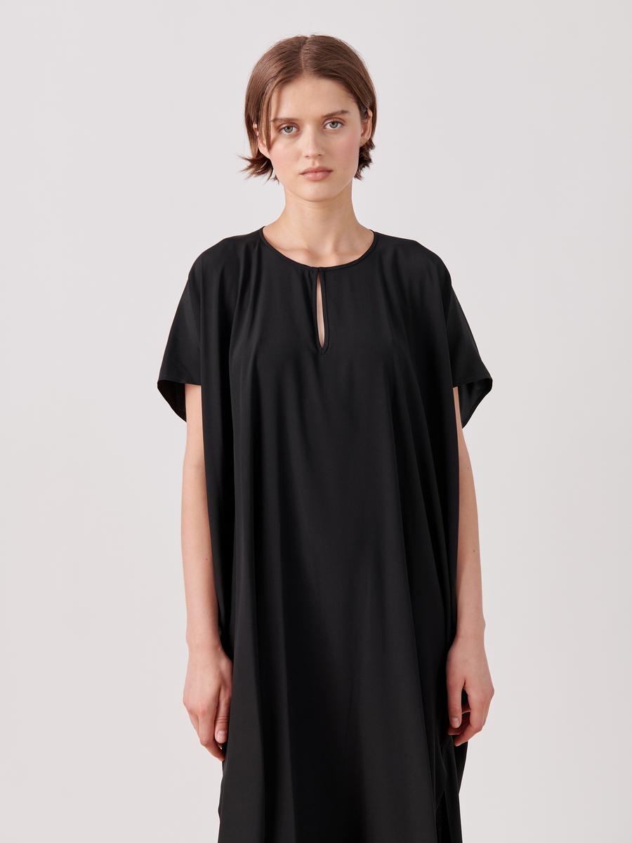 A person with short brown hair wearing a black, mid-length Curve Rae Caftan by Zero + Maria Cornejo made of stretch silk charmeuse with side slits and a small keyhole cutout at the neckline is standing against a plain white background. The person has a neutral expression and their arms are relaxed at their sides.