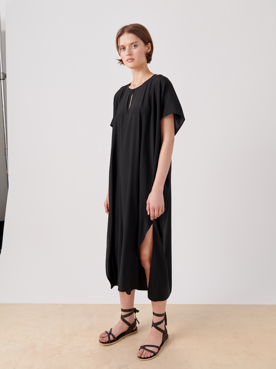 A person is standing against a plain light background wearing a loose-fitting, short-sleeved, black Curve Rae Caftan by Zero + Maria Cornejo with a side slit. They are also wearing black strappy sandals. Their hands are hanging by their sides and they have a neutral expression.