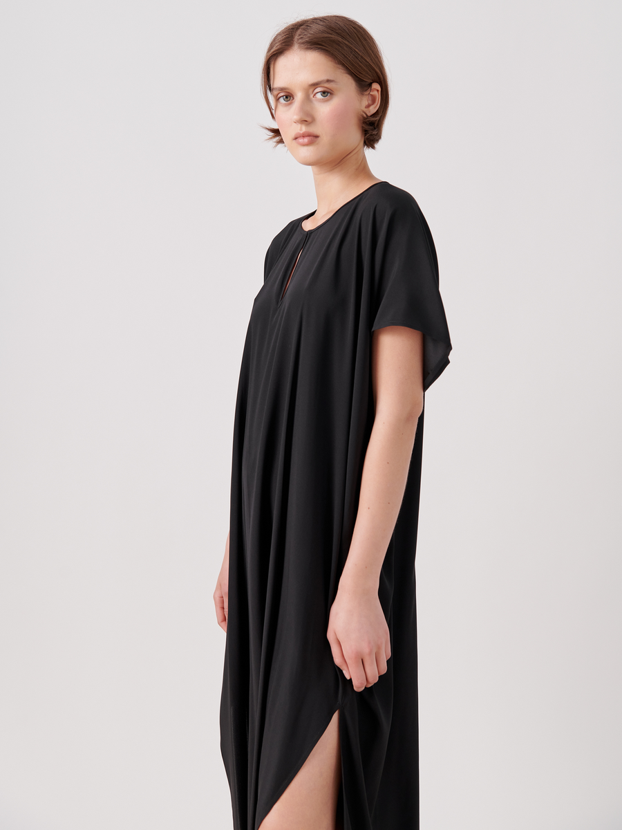 A person with short hair wearing a loose-fitting, black Curve Rae Caftan by Zero + Maria Cornejo stands with a neutral expression. The mid-length dress has a round neckline and short sleeves, with fabric draping elegantly and featuring side slits. The background is plain and light-colored.