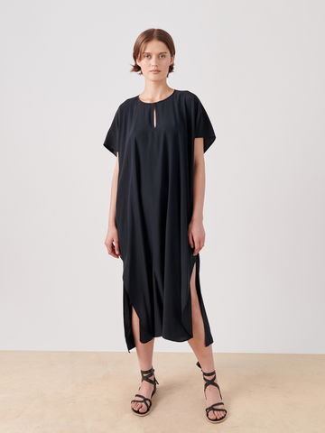 A person stands against a white backdrop wearing the Curve Rae Caftan - Final Sale by Zero + Maria Cornejo, a loose-fitting black silk charmeuse dress with short sleeves and side slits. They pair this elegant mid-length dress with black strappy sandals.