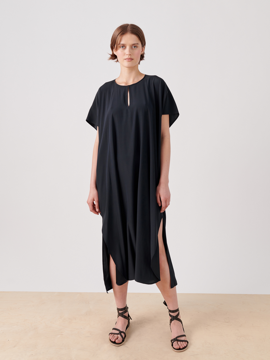 A person stands against a plain white background, wearing the Curve Rae Caftan by Zero + Maria Cornejo. The loose-fitting black dress with short sleeves and high side slits features a small keyhole cutout at the neckline. They are also wearing black strappy flat sandals.