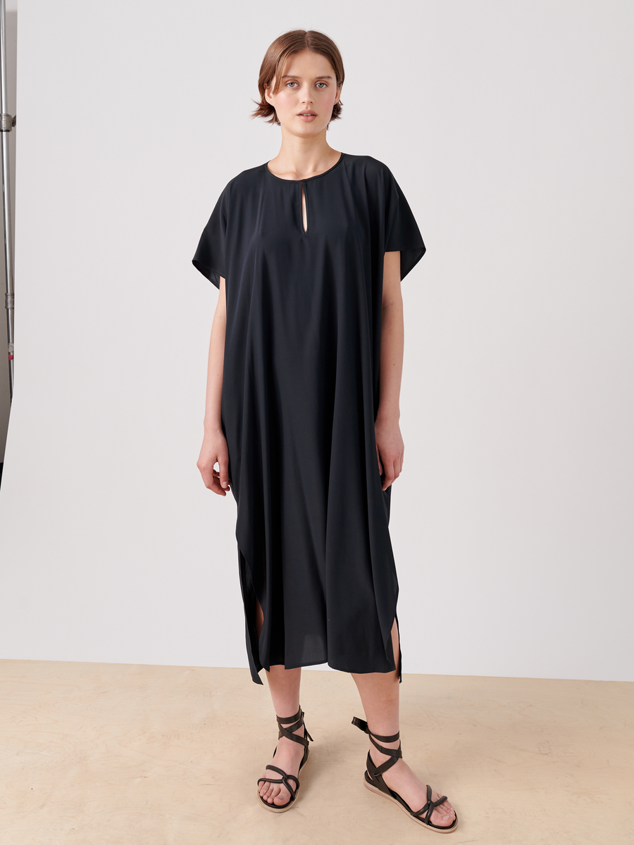 A person with short hair stands against a plain background, wearing a loose-fitting, mid-length black Curve Rae Caftan by Zero + Maria Cornejo with short sleeves and small side slits. They have on black strappy sandals, and their posture is relaxed with arms at their sides.