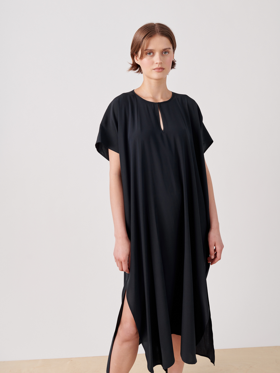 A person with short brown hair is wearing a loose-fitting, black Curve Rae Caftan by Zero + Maria Cornejo with a small keyhole neckline and short sleeves. The mid-length dress has side slits and drapes down asymmetrically. They are standing indoors against a plain white background.