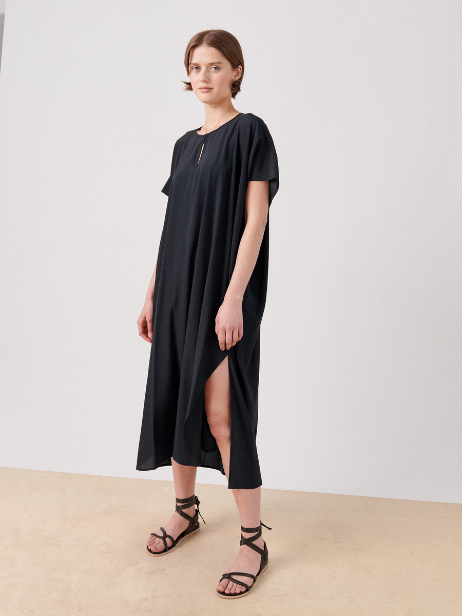 A person stands against a plain background wearing the Curve Rae Caftan by Zero + Maria Cornejo, a loose-fitting, mid-length black dress with short sleeves and a side slit. They have short hair and are wearing black lace-up sandals.
