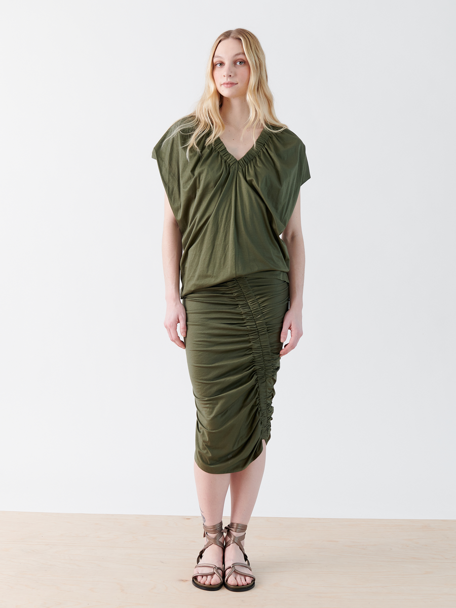 A woman stands on a light wooden floor against a white background, wearing a sustainable dark green set with short sleeves and the Kia Skirt from Zero + Maria Cornejo. She pairs it with strappy brown sandals, her long blonde hair flowing freely as she maintains a calm expression.