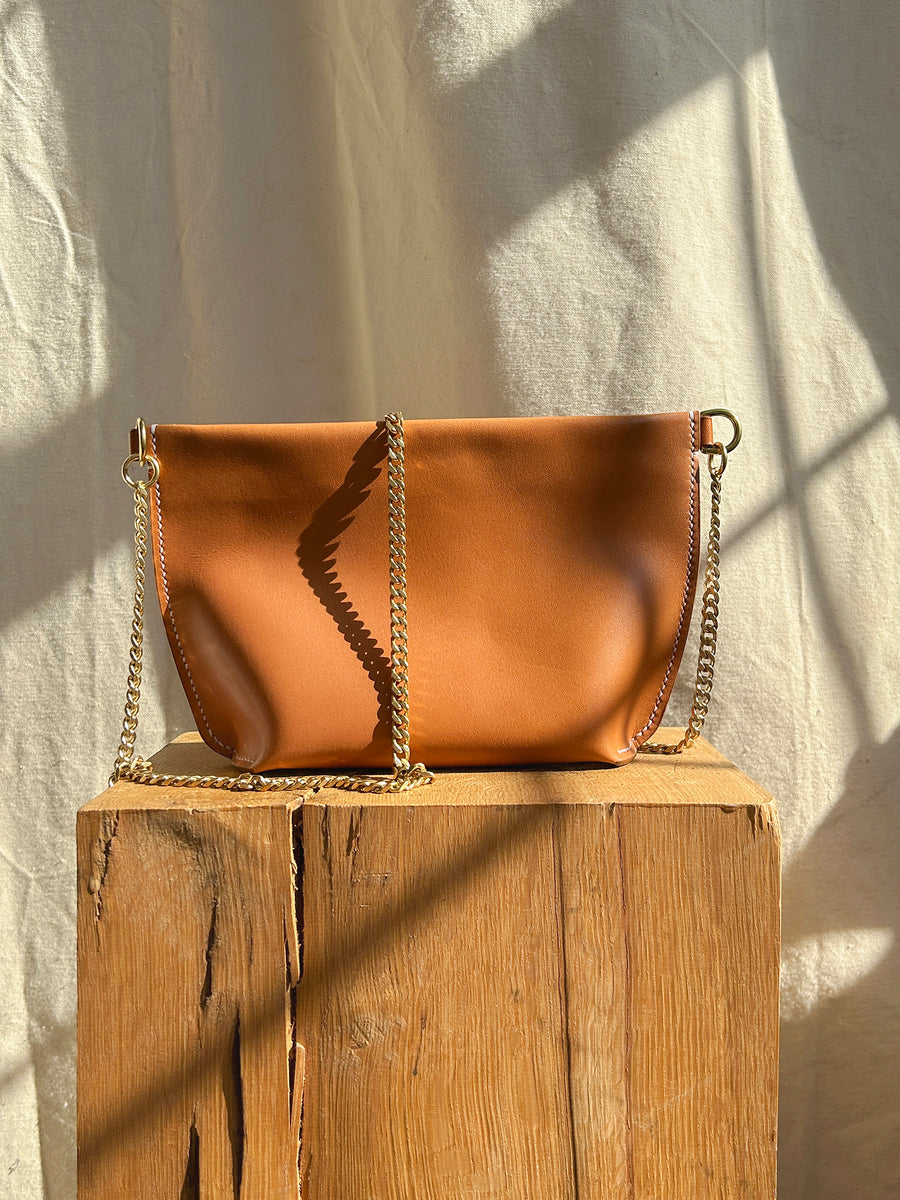 The Bartleby Objects Stowe Chain Bag, crafted from brown leather with gold chain straps, and hand-made in New York City, is showcased on a wooden block. Sunlight creates shadows on the textured backdrop, accentuating the bag's smooth surface and wavy seam detail.