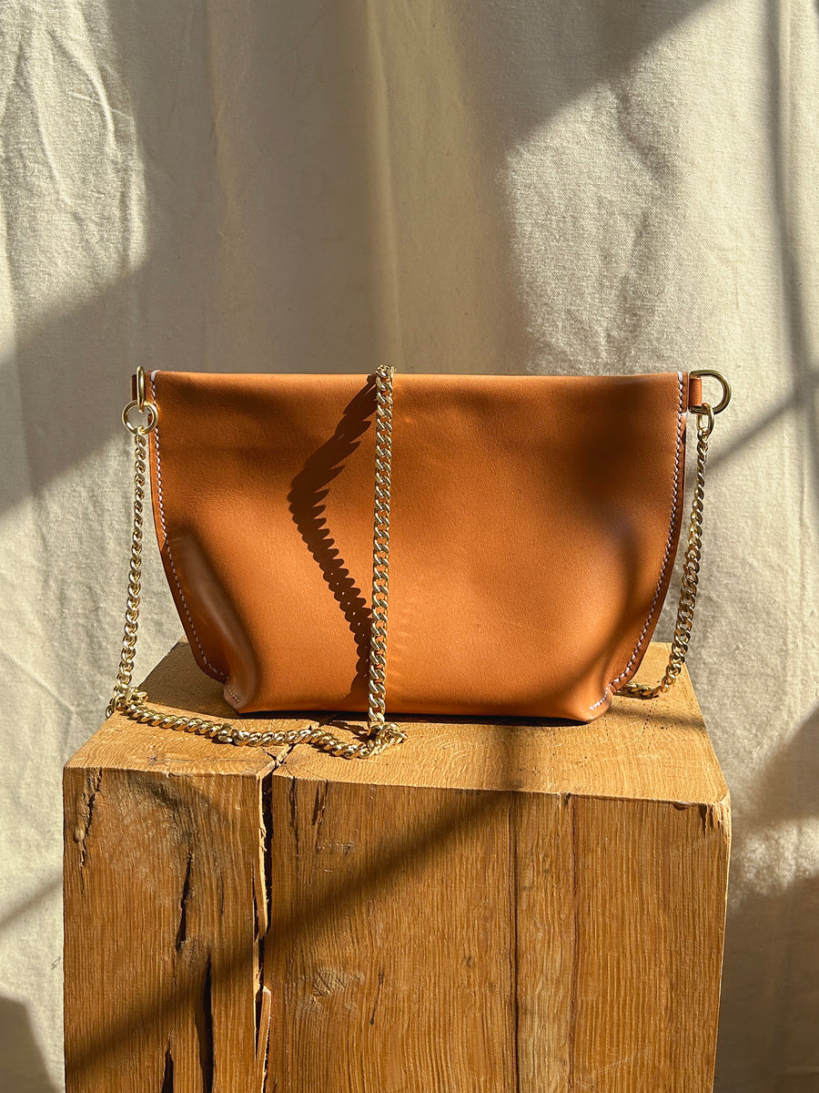 The Bartleby Objects Stowe Chain Bag, crafted in tan leather and featuring a solid brass chain strap, rests on a wooden platform as warm sunlight casts shadows on the cream-colored backdrop.