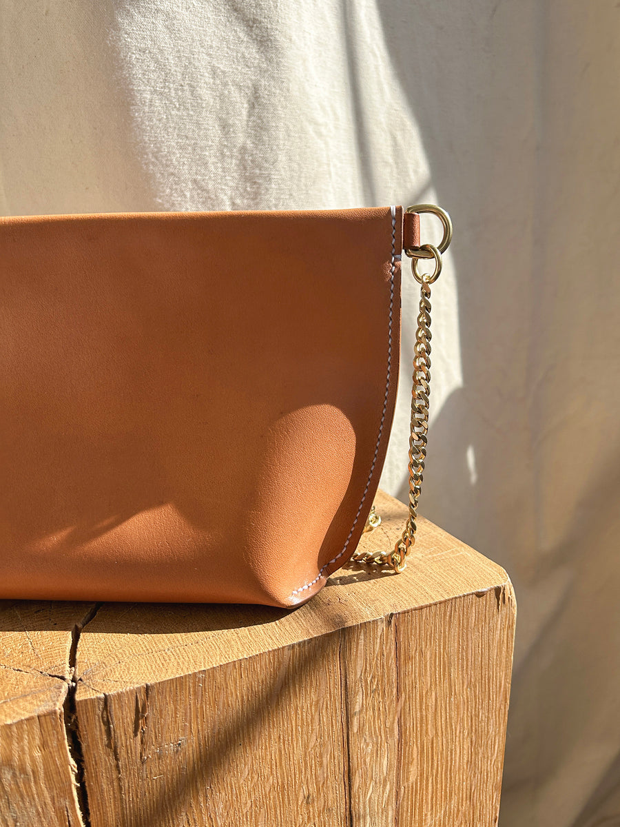 Close-up of the Bartleby Objects Stowe Chain Bag, showcasing its brown saddle-stitched design and solid brass chain strap as it rests on a wooden surface. The background features a soft, off-white fabric that complements the scene, while the lighting accentuates the texture of its vegetable-tanned leather.